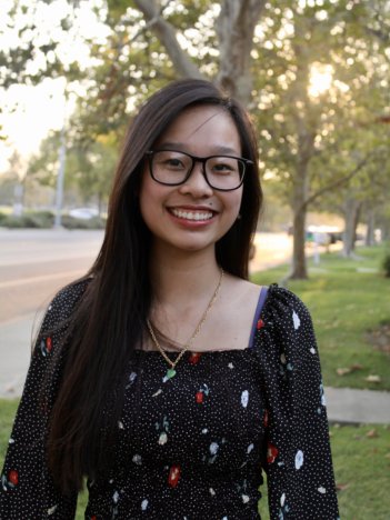 A young asian woman with long dark brown hair and black-rimmed glasses wears a black dress with a white and red pattern and smiles at the camera in front of a street.